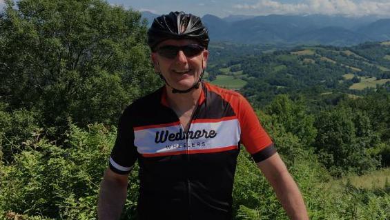Tim Blackburn is stood with a bike in front of him. He is wearing a lycra top, cycling gloves, helmet and sunglasses. He is looking at the camera and smiling. The landscape behind him is green, full of trees and there are hills in the distance.