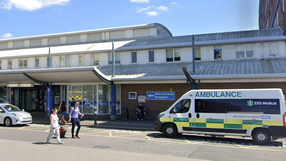The outside of the North Tees Hospital Trust reception in the sun. An ambulance is parked outside the building and a sign on the wall reads 'Main entrance reception' with an arrow.
