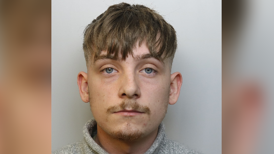 A mugshot of Taylor Stacey. He is wearing a grey jumper and has short mousey brown hair with blue eyes and a solemn look on his face.