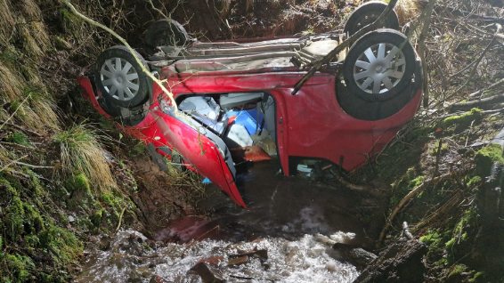 An overturned red car is balanced above a stream on two banks with vegetation on. The door is left open.