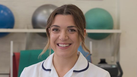 Chloe Boobier dressed in her white physio uniform and is smiling for the camera. She has long brown hair that has been tied back.
