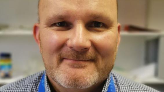 A man wearing a blue checked shirt in with an office backdrop behind him.