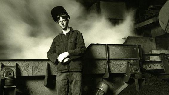 A black and white photograph of a foundry worker leaning on a piece of machinery. The man looks quite young and is wearing a safety visor on his head which is tipped up, revealing his face. The background appears to be smoky.