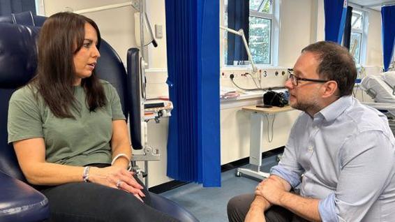 Emma Cullen sat on a hospital char, chatting to a male doctor who is wearing a blue shirt and glasses and is kneeling down in front of Emma