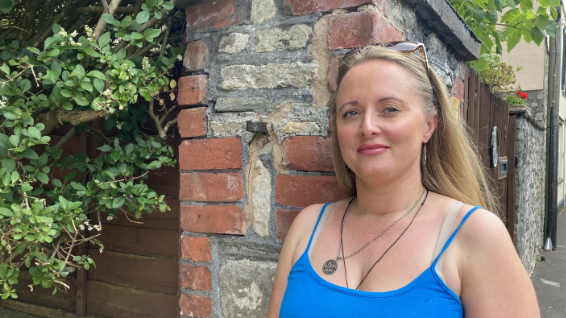 Kelly Sheppard wearing a blue top stood next to the wall with carvings written on. 