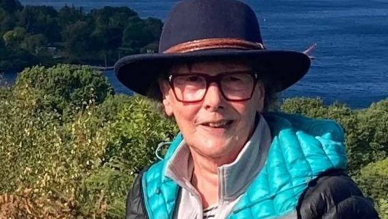 Sally Hall smiles wearing a wide-brimmed hat and a waterproof coat on a hill overlooking a lake. 