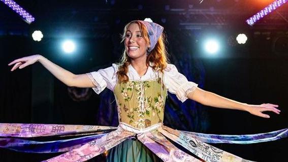 A red-haired Ellana Gilbert on stage wearing a white top and a green vintage-style corset. There are multi-coloured strips of material strung out from her waist