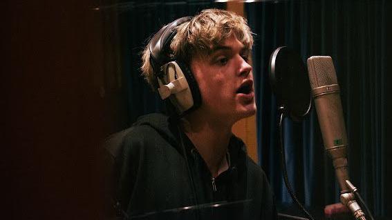 Oscar in a music studio singing in front of a microphone. He has headphones on and has short blonde hair and a black jacket on. 