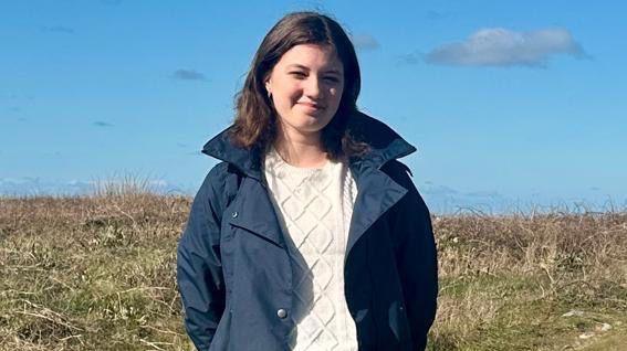 A picture of Eva stood in a field. She is wearing a navy coat and is surrounded by grass.