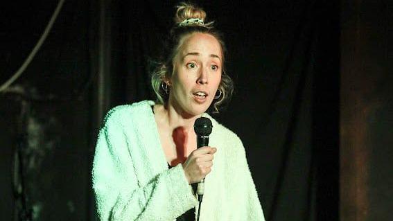 A blonde woman in a mint green dressing gown stands on a dark stage holding a microphone.