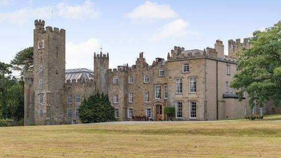 A large property with a castellated roof and a tower at the front with a clock.
There is a large expanse of lawn in front. There are also a number of mature trees to either side. A driveway can be seen coming from the right hand side and leading to the front of the property.