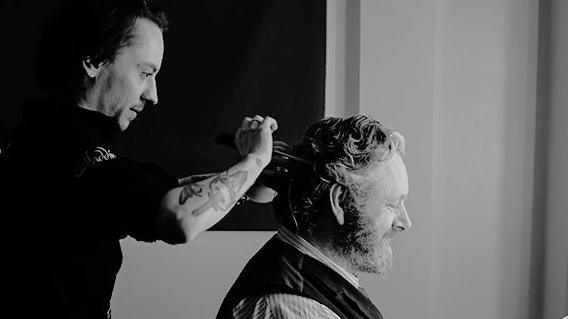 a black and white photograph of actor Michael Sheen, who is sat facing away from the camera, wearing a striped shirt and waistcoat. A man with a tattooed arm is stood behind him, styling his hair.