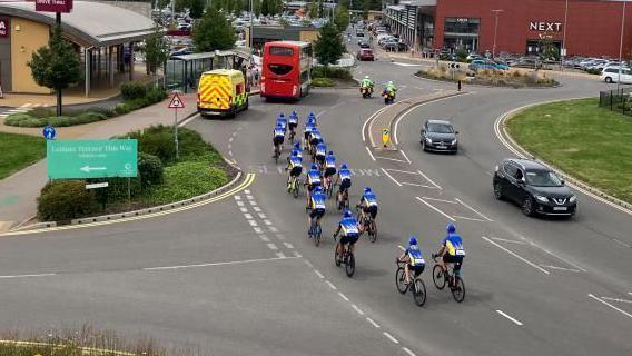 A peloton of bikes with riders in blue and white tops on a road next to Rushden Lakes