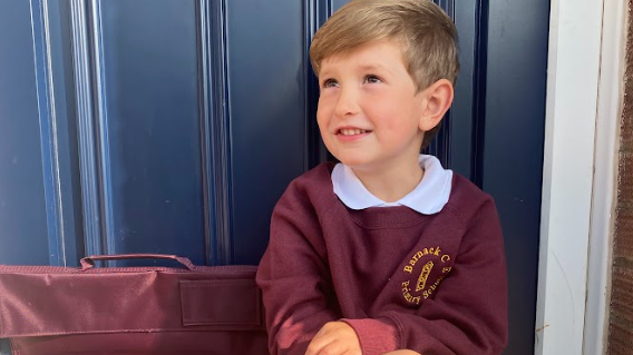 Benedict sits on his front step in front of the navy blue door. He wears a maroon logo sweater and his matching briefcase bag is next to him. He is looking upwards and smiling at someone to the left of the photo.