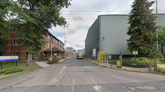 An entrance to a factory with a red brick, three-storey, building on the right and the grey factory on the left. 