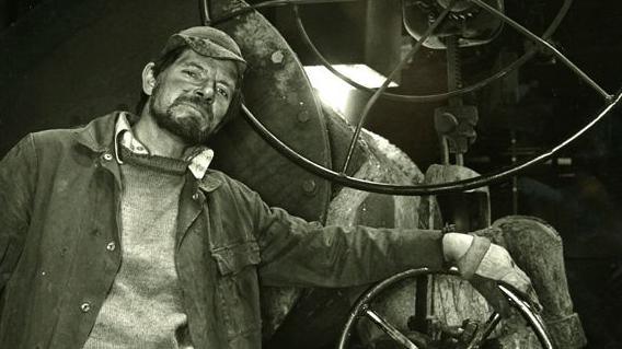 A black and white photograph of a foundry worker posing alongside some machinery. He has a full beard and is wearing a cloth cap and jumper underneath unbuttoned overalls. He is looking at the camera and leaning on a large wheel which is attached to the machine.