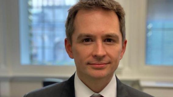 Mark Sewards wearing a grey suit, white shirt and red tie staring into the camera