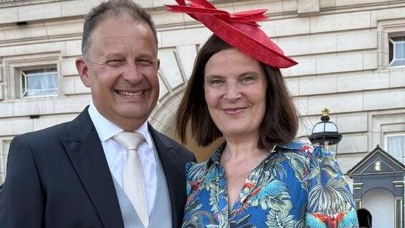 A man and woman stand side-by-side outside Buckingham Palace. The man wears a black suit, light blue waistcoat and a light tie. The woman has a blue shirt dress with a green and red floral pattern and a red hat.