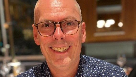 A man wearing a navy floral top and round brown glasses. The background of a restaurant is blurred.