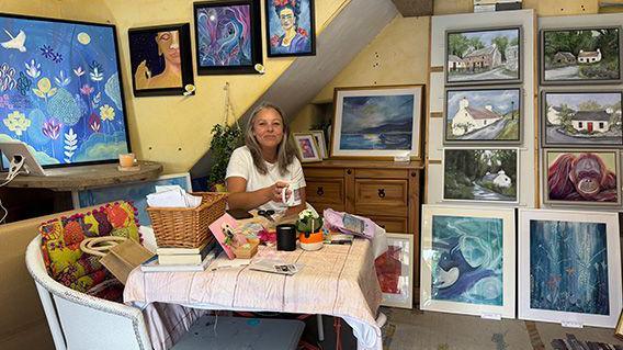 A woman sitting at a table in a small venue holding a mug. She has long blonde hair and is wearing a white T-shirt and is surrounded by paintings which area a mix of landscapes and portraits.