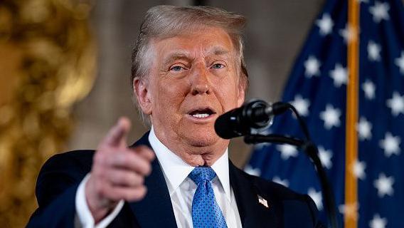 Trump at microphone with blue tie and dark suit before an American flag 