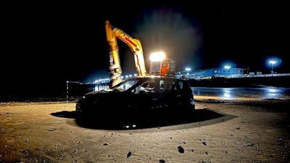 A black car stuck on a beach being rescued by a recovery vehicle at low tide in the dark