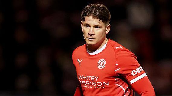 Max Sanders wearing his red Crewe home shirt and is looking down to his right at the ball