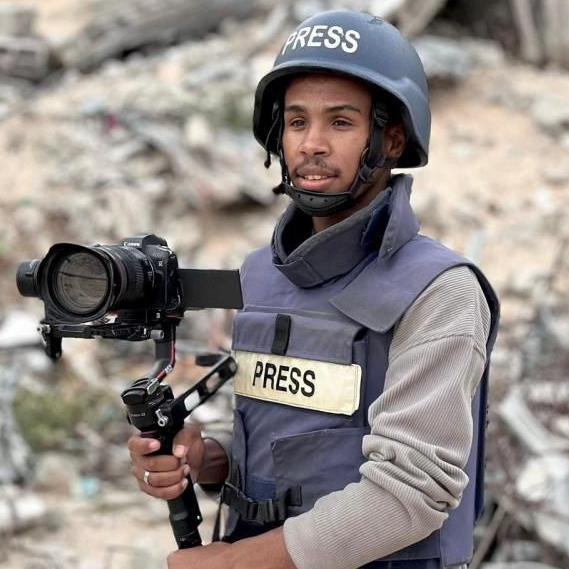 A man wears a press vest holding a camera and wears a helmet. He is stood infront of rubble