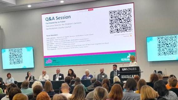 A meeting with a top table of panellists all wearing badges and sitting in front of a projected screen titled "Q&A session". A woman with long brown hair is standing at a lectern. There are QR codes on screens either side of the main projection. Rows of audience members are watching.