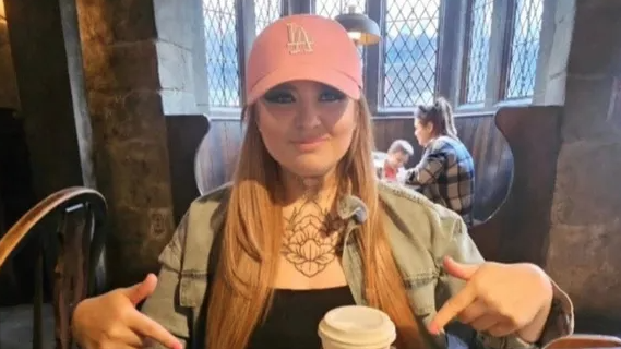 June Bunyan - a woman with long reddish hair past her shoulders and a pink baseball cap - sits inside a pub or a cafe and points to a drink and food in front of her.