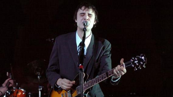 Pete Doherty stands on stage singing into a microphone and playing an electric guitar while performing with The Libertines during The Libertines in Concert at Brixton Academy in London in 2004. He has brown hair and is wearing a black suit with white shirt and black tie. The corner of a drum kit can be seen behind him to the left. The background is black. 