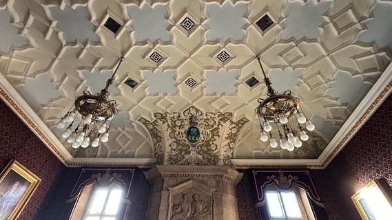 An ornate ceiling, carved and painted in cream and pale blue. Two large chandeliers hang down over tall windows. There is also an engraved tablet.