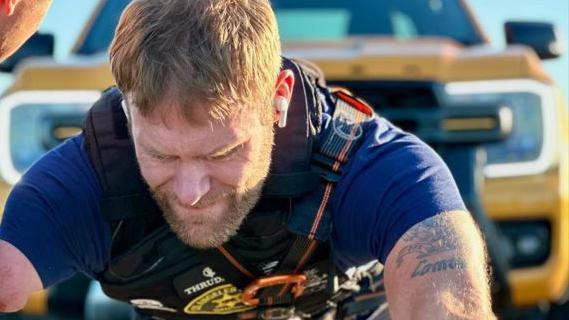 Mark Ormrod MBE is seen looking downwards with gritted teeth with a large yellow vehicle in the background