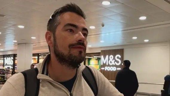 Një burrë i ri me mjekër dhe në çantë shpine qëndron në një sallë aeroporti.