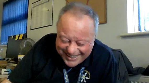 Dave Kelly, wearing a black top, laughs and smiles as he is photographed in front of a desk. 