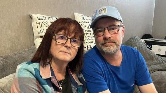 Louise, wearing a blue checkered shirt and glasses, sits next to Clint Slater, wearing a Coventry City cap, glasses and a blue T-shirt, on a grey sofa. Behind them are two white cushions with different coloured lettering on them spelling our words - we can only see some like "when you miss".