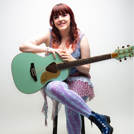 Dynes ifanc gyda gwallt coch hir. Mae'n eistedd ar stôl gyda gitar gwyrdd yn ei chôl. 