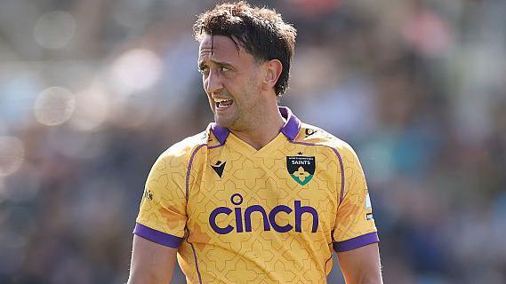 Northampton Saints rugby union player Alex Mitchell looks to his right while playing a match, wearing a yellow shirt with purple trim