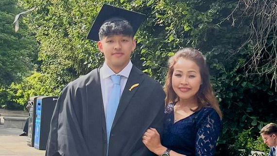 A young man wearing a university graduation gown. His mother is stood on his left. They are stood outside in front of some trees and looking at the camera