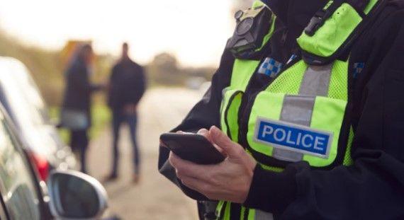 A generic photo of a policeman taking down a driver's details by the side of a road, while two blurred figures stand in the background.