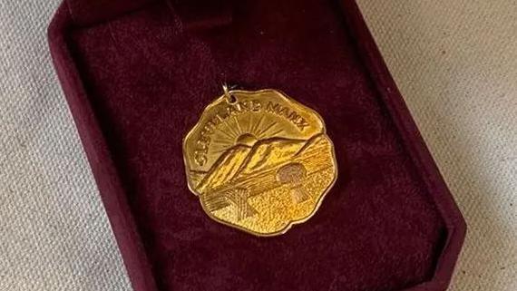 A gold medal in a burgundy velvet box. The medal is engraved with a scene featuring a field with wheat sheafs standing in it in front of the sun rising behind hills, with the word Cleveland Manx above it.
