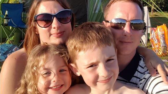 Louise Ryan and her late husband Nick, with their two children in front of them smiling.