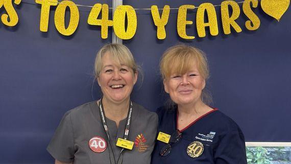 Two nurses with blonde hair stand smiling side by side under a banner which celebrates their 40 years in the job.