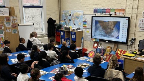 A school classroom with children stat at desks looking at a television mounted on the wall