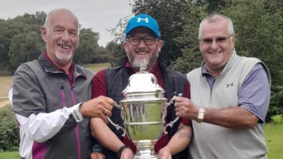 Three men smile at the camera as they hold a large silver trophy in front of them.