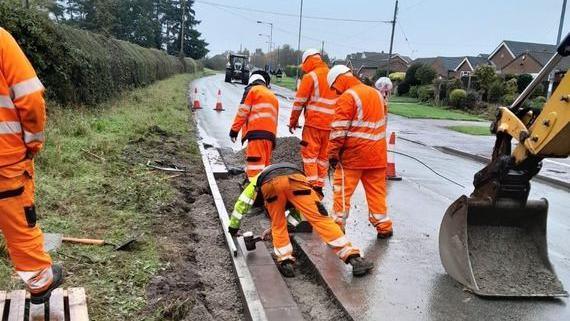 Workers in bright orange overalls repair a section of road. There is a digger bucket in the right hand side of the image and a tractor in the distance. Houses can be seen on the right side of the image, next to the road.