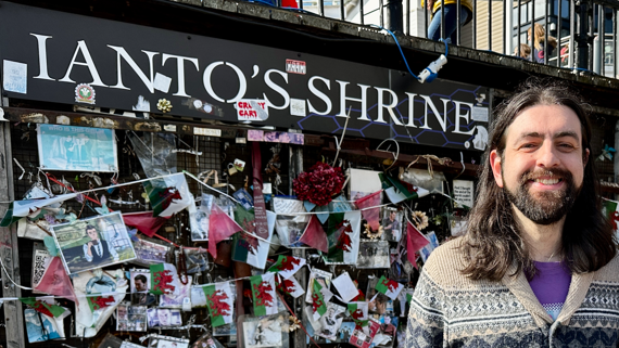 A wall with the words Ianto's Shrine and hundreds of photographs and Welsh flags pinned to it. In front of it is a man with long black hair and a beard, wearing an Aztec print cardigan and smiling at the camera.