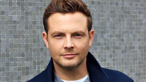 Mark Rhodes, a man with short brown slightly wavy hair is slightly smiling - he has a bit of stubble on his chin and is wearing a grey jumper and navy blue jacket