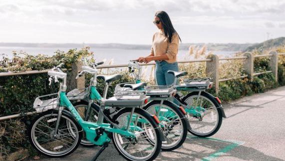 Three light green bikes standing up aside a path overlooking the coast, with a woman with sunglasses stood next to one, trying to get it started 