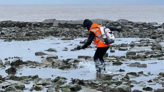 A person wearing a black hoodie and orange high-vis vest is wading through rock pools by a beach with the grey sky and sea behind them.